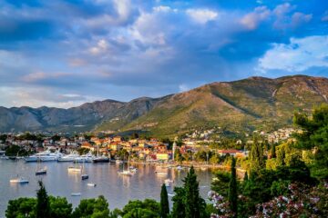 boats on body of water surrounded by trees and houses near mountain under blue and white sky at daytime